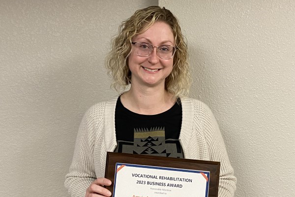 Woman with medium length blonde curly hair, wearing glasses, black shirt, and a beige cardigan. She is holding an award.