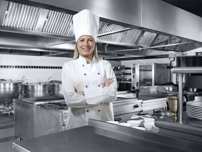 A female chef wearing a white chef uniform and standing in commercial kitchen, while smiling with crossed arms