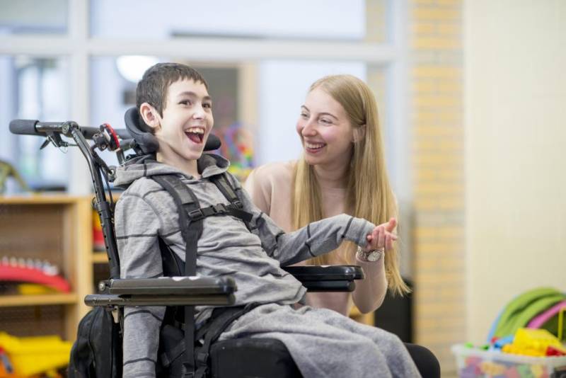 Young boy in a wheelchair with and adult smiling at his side. 
