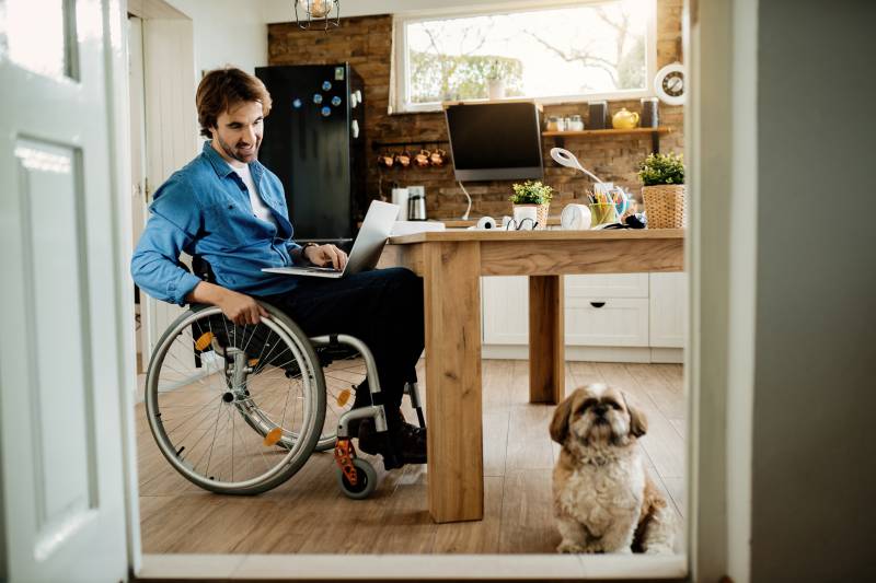 Man in wheelchair, using a computer in his kitchen, with his small pet dog