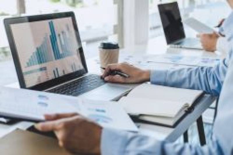 Someone sitting at a desk looking at a computer with data and holding a piece of paper with data.
