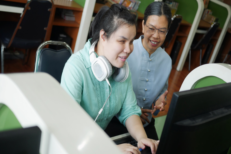 Person that is blind working on the computer with an instructor next to her. She is wearing white headphones around her neck.