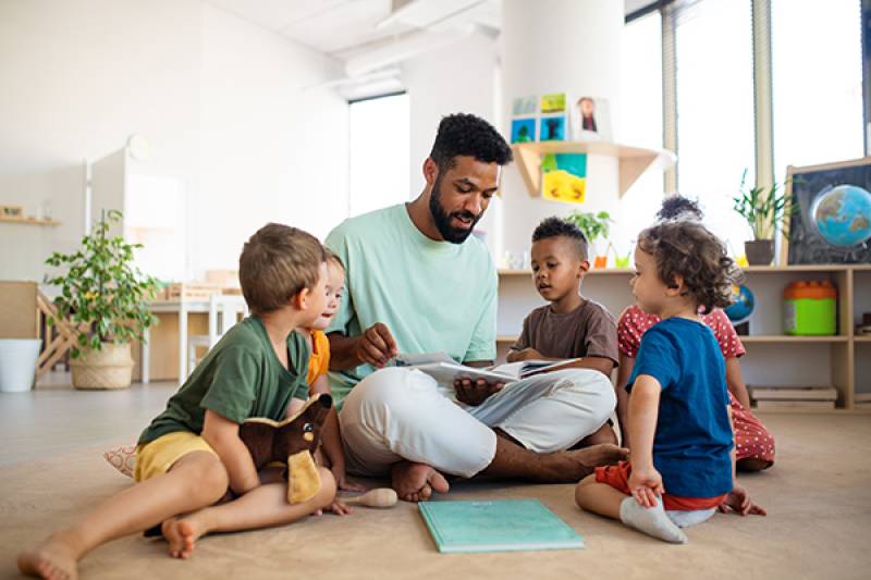 Group of small nursery school children with man teacher sitting on floor indoors in classroom, having lesson.