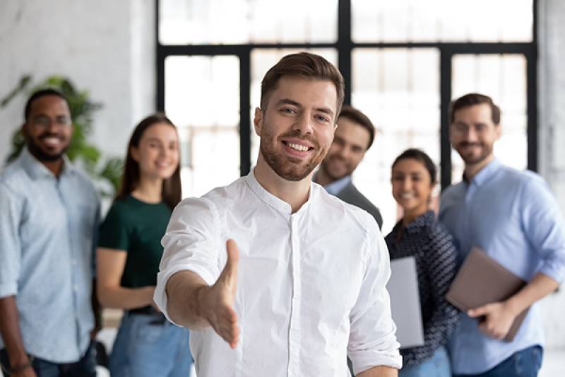 Smiling Caucasian businessman stretching hand to shake hands. There are other office workers in the background.