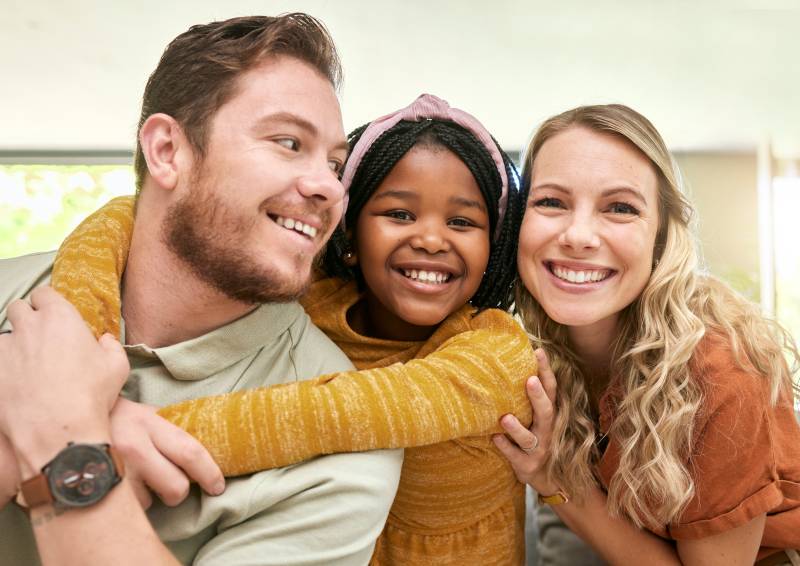A child in a yellow sweater hugs two adults from behind. Everyone is smiling warmly, conveying happiness and closeness