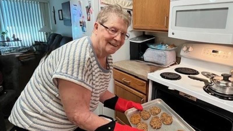 Older woman with short gray hair and wearing glasses getting cookies out of the oven.