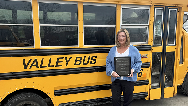 Middle aged female with medium length blonde hair and glasses holding an award in front of a yellow school bus.