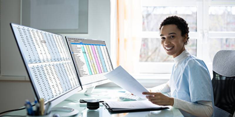 Worker working on two computers with paperwork on her desk.