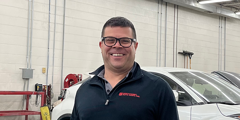 A light skinned middle-aged male with short brown hair, glasses, a black quarter zip shirt, and khakis. He is standing in the garage of the auto body shop next to a black truck with the passenger door open.