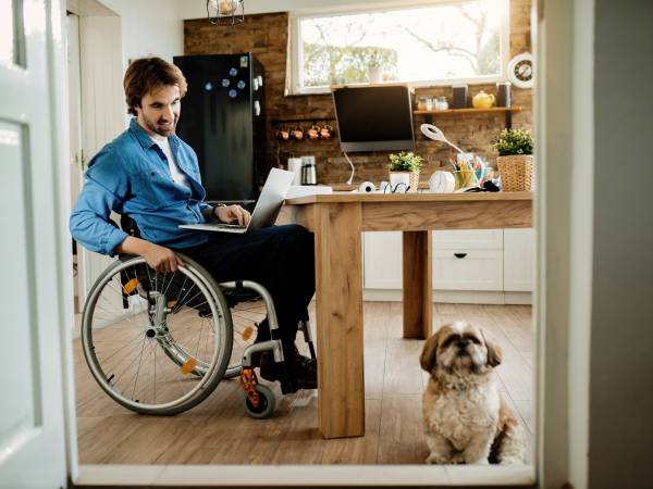 Man in wheelchair, using a computer in his kitchen, with his small pet dog