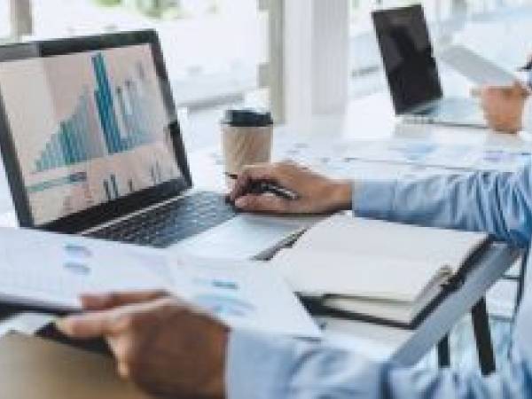 Someone sitting at a desk looking at a computer with data and holding a piece of paper with data.