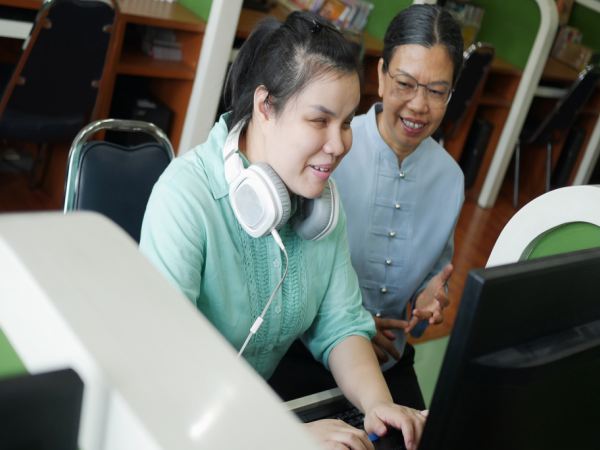 Person that is blind working on the computer with an instructor next to her. She is wearing white headphones around her neck.