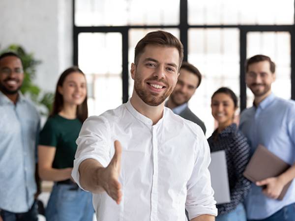 Smiling Caucasian businessman stretching hand to shake hands. There are other office workers in the background.