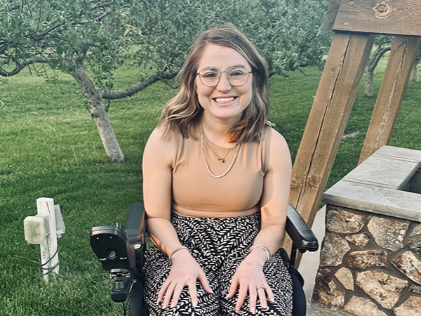 Female with medium brown hair, glasses and a tan tank top sitting in a wheelchair outside.