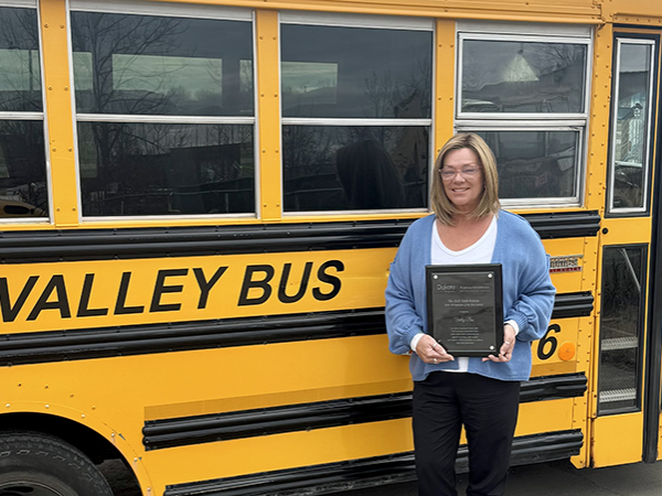 Woman standing by school bus holding award.