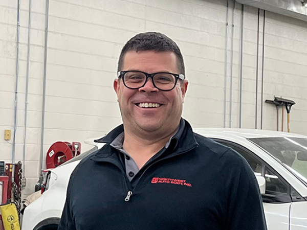 A light skinned middle-aged male with short brown hair, glasses, a black quarter zip shirt, and khakis. He is standing in the garage of the auto body shop next to a black truck with the passenger door open.