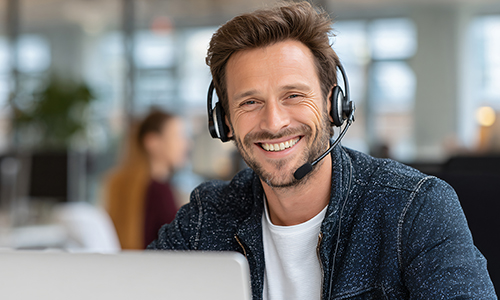 Man wearing headset with microphone, smiling while working on laptop. Technical support or customer service concept.