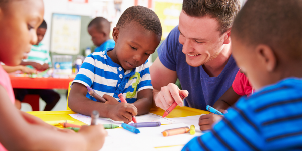 young children coloring at table with male teacher