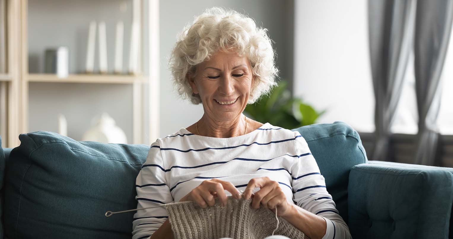 smiling older woman sitting on a blue couch knitting