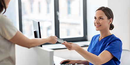 A medical professional, wearing scrubs, sitting at her desk, taking a card from someone.