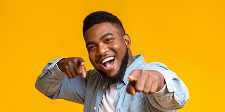 A man, against a solid orange background, smiling and enthusiastically pointing at you with both hands.