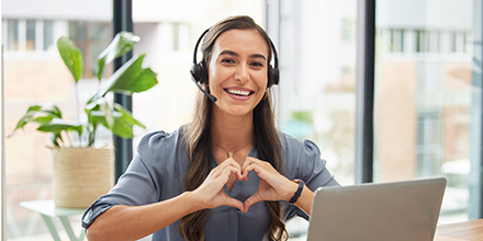 A woman sitting in front of her computer, wearing headphones, and making a heart shape with her hands.