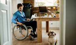 Man in wheelchair, using a computer in his kitchen, with his small pet dog