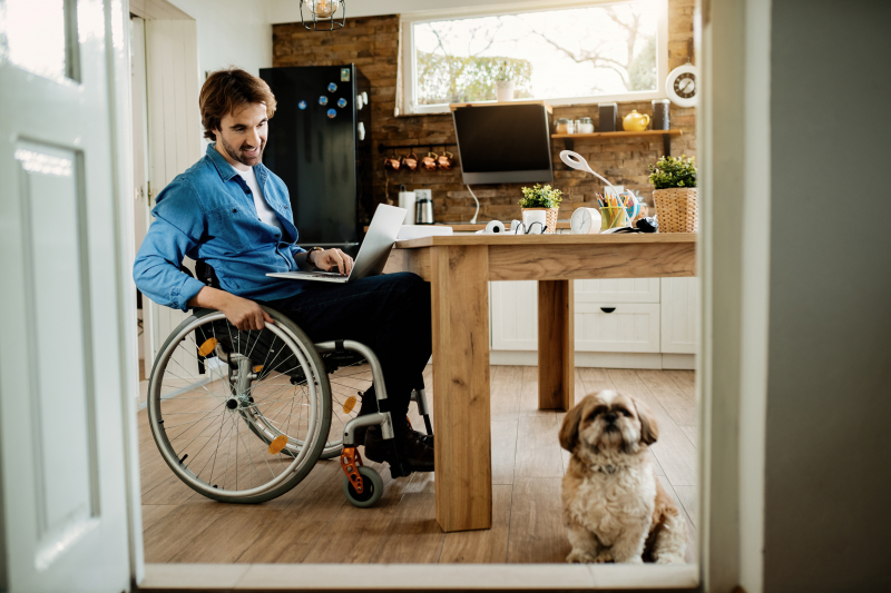 Man in wheelchair, using a computer in his kitchen, with his small pet dog